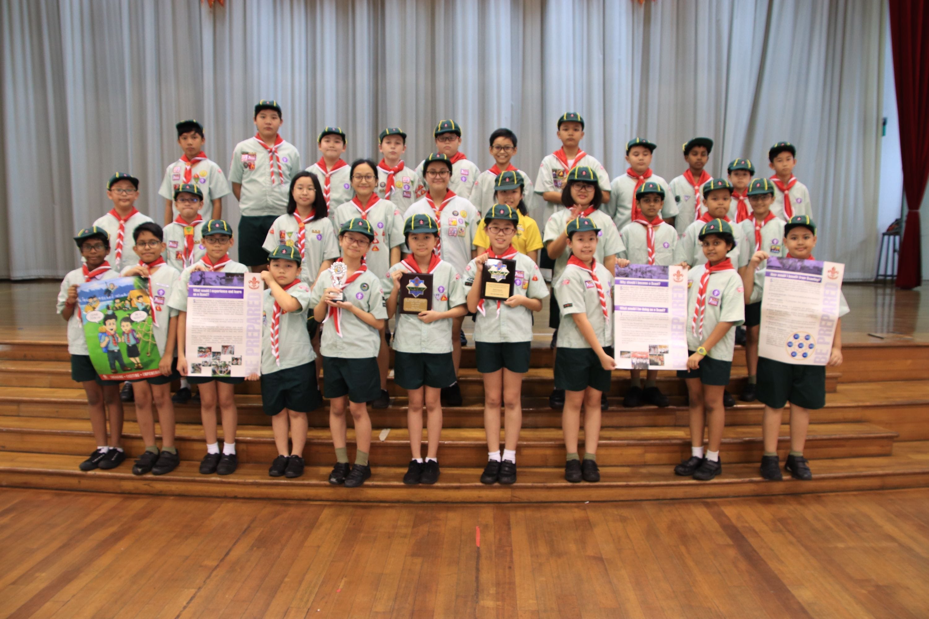 Group of young scouts in green uniforms and red neckerchiefs posing on stage steps, holding trophies and poster boards.
